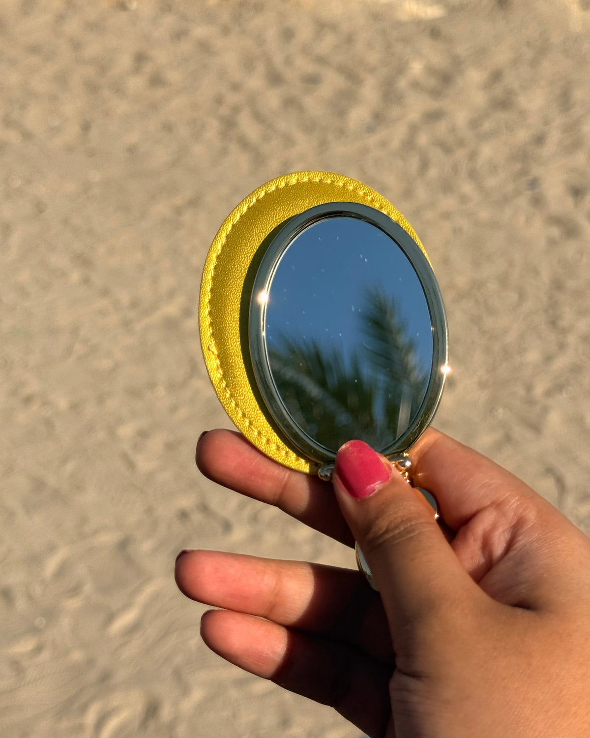 A hand with pink nail polish holds a small oval mirror with a bright yellow textured frame, reflecting palm trees against a sandy beach background.