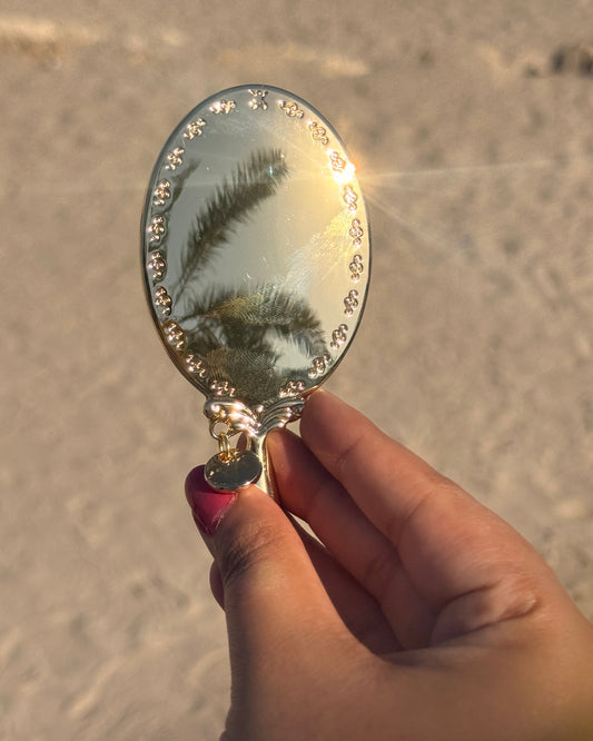 A hand holding a small oval hand mirror with a silver frame, reflecting palm trees against a bright sunlit sandy beach background.